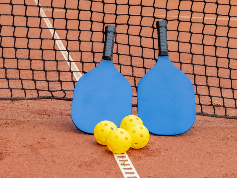 Hai cây vợt pickleball xanh dương và ba quả bóng vàng đặt cạnh lưới