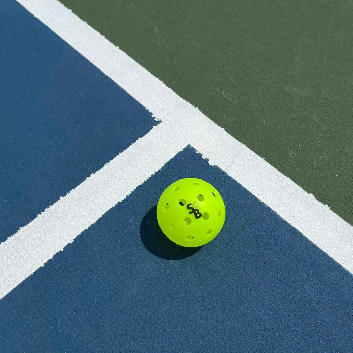 Quả pickleball màu vàng chanh nằm cạnh vạch kẻ trên mặt sân xanh
